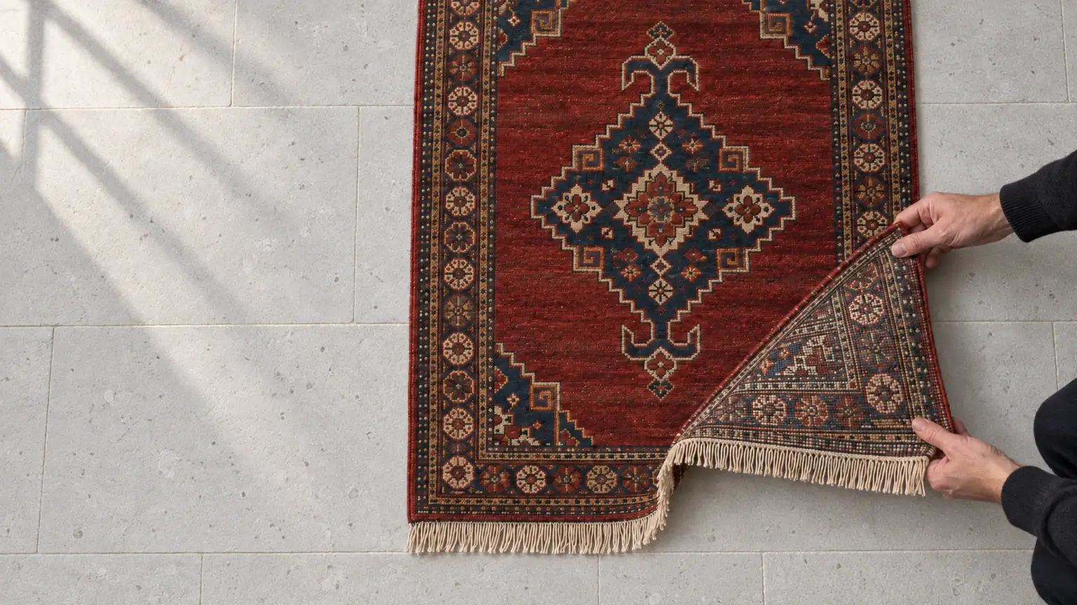 Two hands turning back the corner of a rug on pale limestone, showing the grid of knots on its back