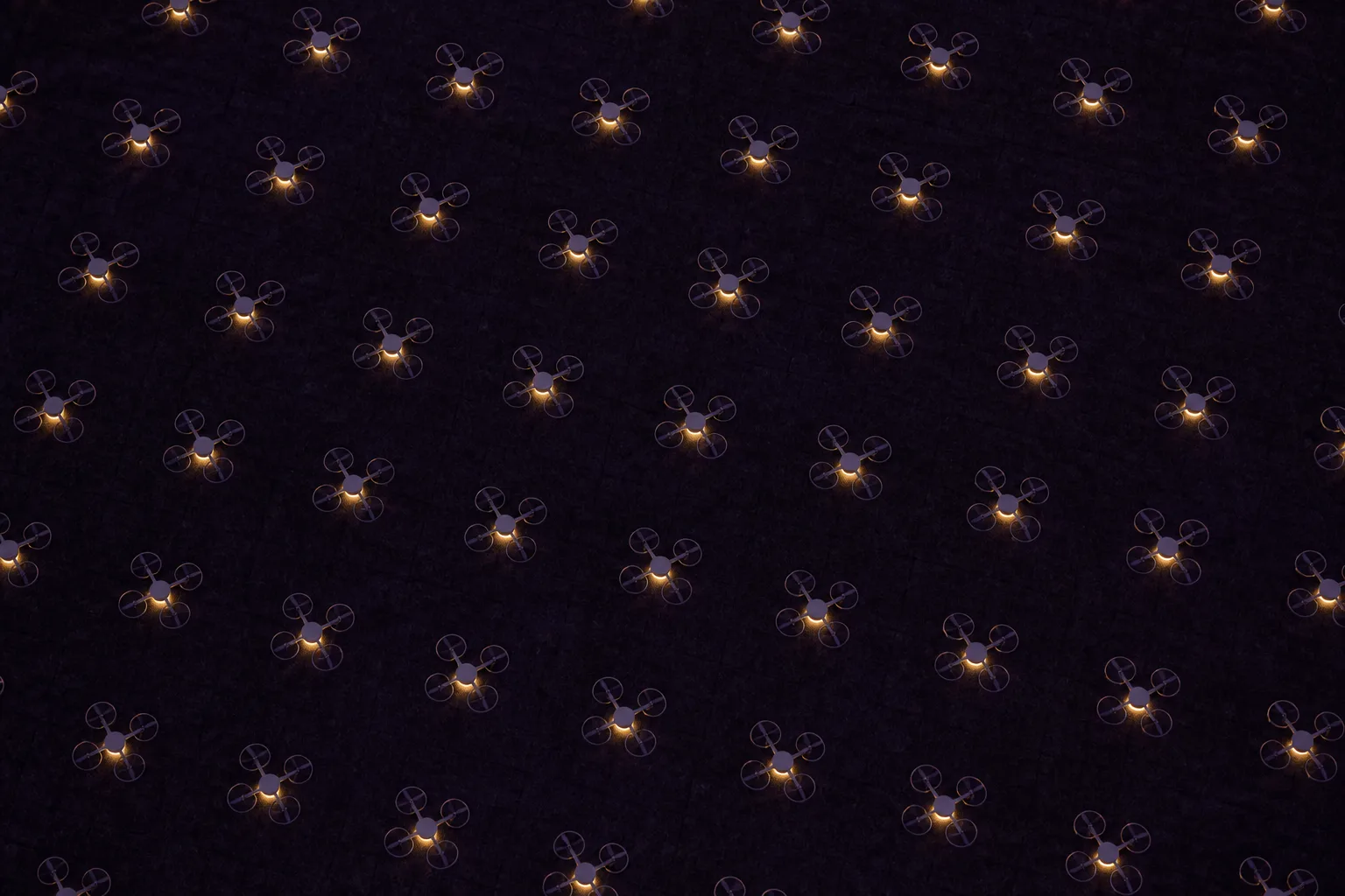 Rows of small drones laid out in a grid on dark grass at dusk, seen from directly above, each with a dim warm light.