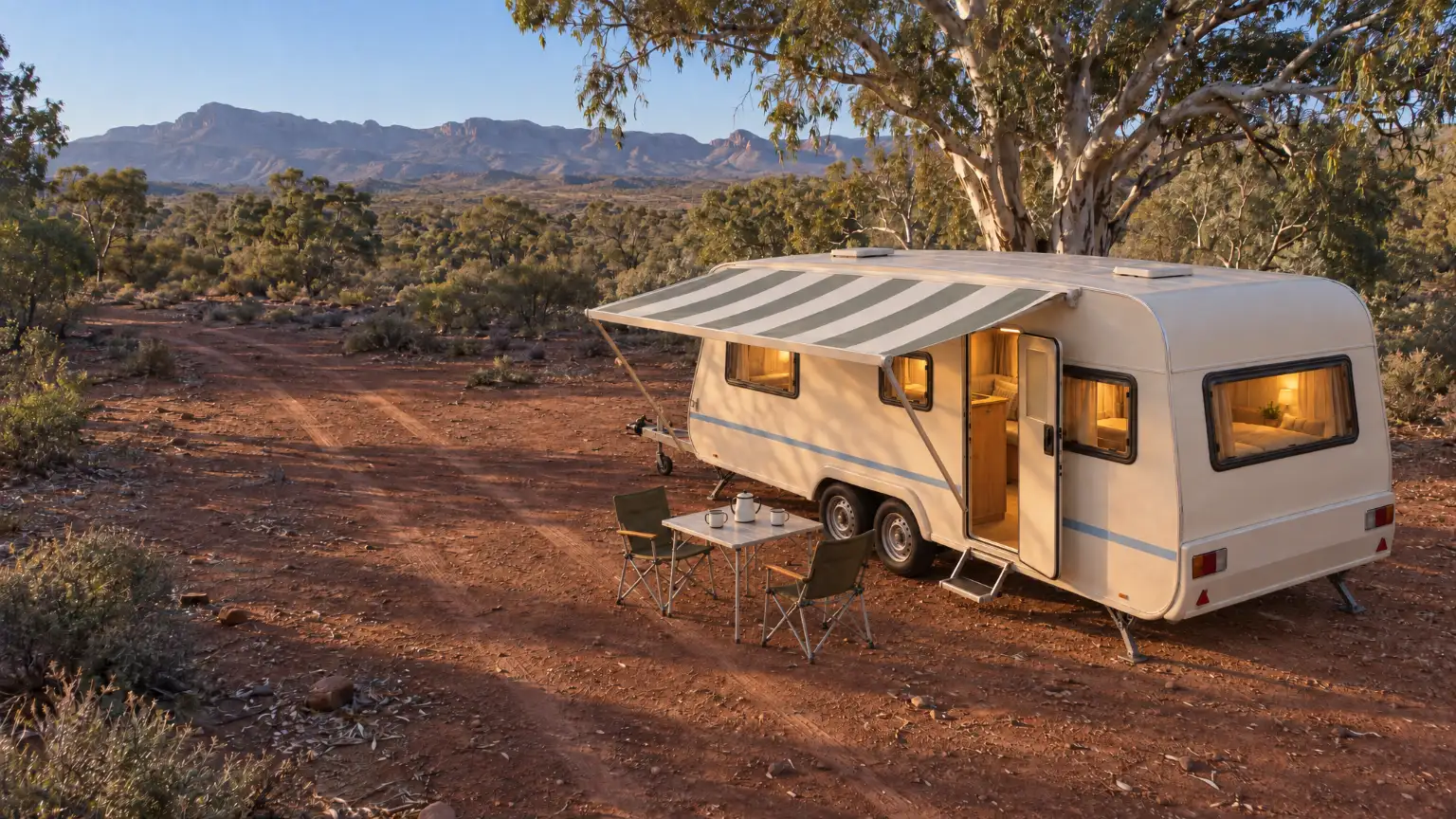 A cream caravan set up under a river red gum on red earth, its grey-green striped awning out, step down and warm lamps on inside.