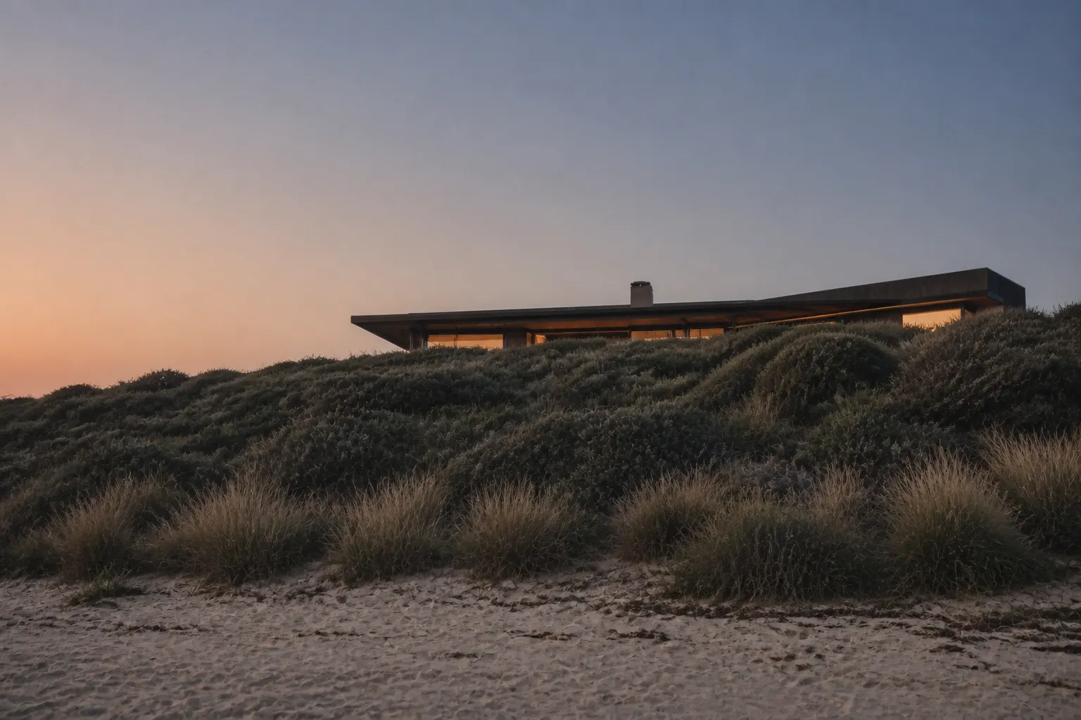 The Aldinga dune house seen from the beach at dusk, its low charred-timber roofline just showing above the crest of the dune