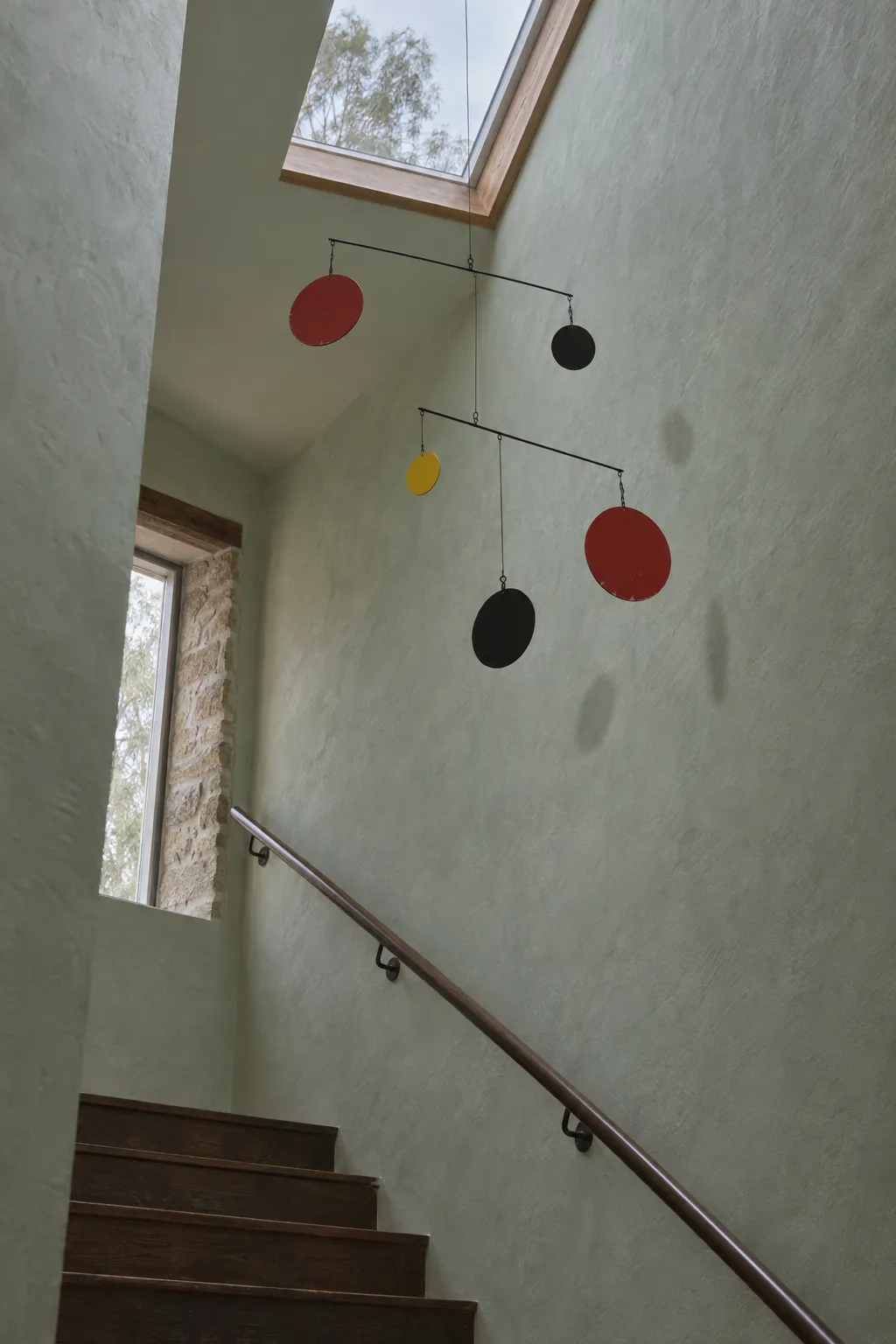 A small hand-made mobile of black rods and vermilion, black and yellow steel discs hanging in a pale stairwell, its shadows soft on the plaster.