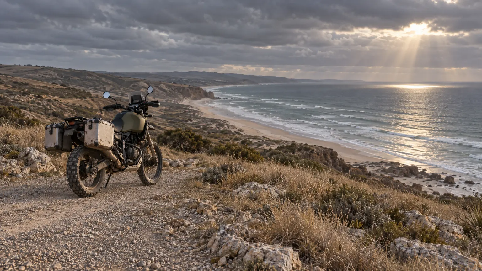 The same adventure motorcycle parked at a cliff-top gravel lookout above a long beach and a grey sea.