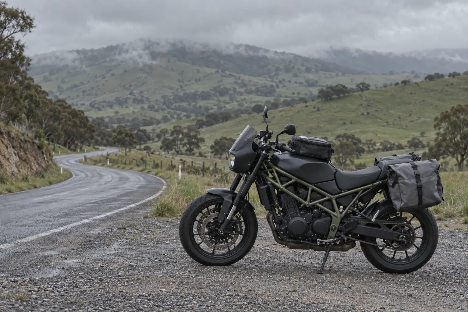 A touring motorcycle with soft panniers parked at a gravel pull-out beside a damp country road in green hills.