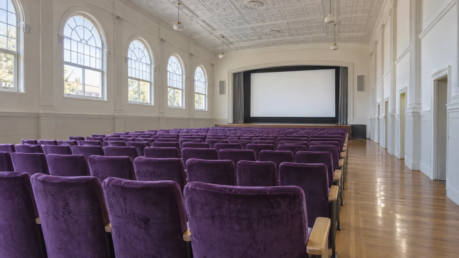 The big room in daylight with the house lights up: rows of orchid velvet seats facing a white screen under tall arched windows