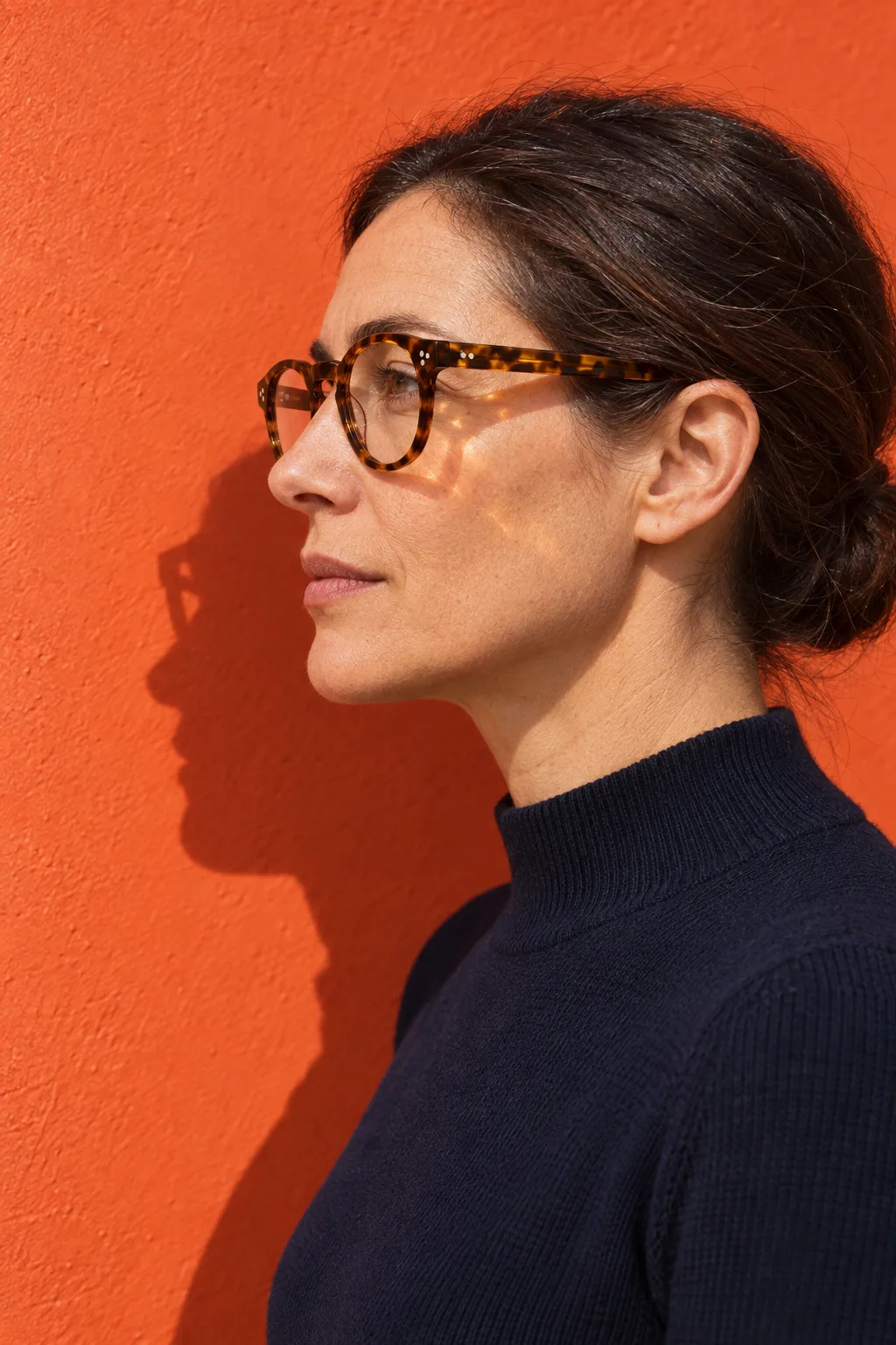 A woman in profile wearing a tortoiseshell panto frame, its temple running straight back over her ear, against an orange wall in hard sun