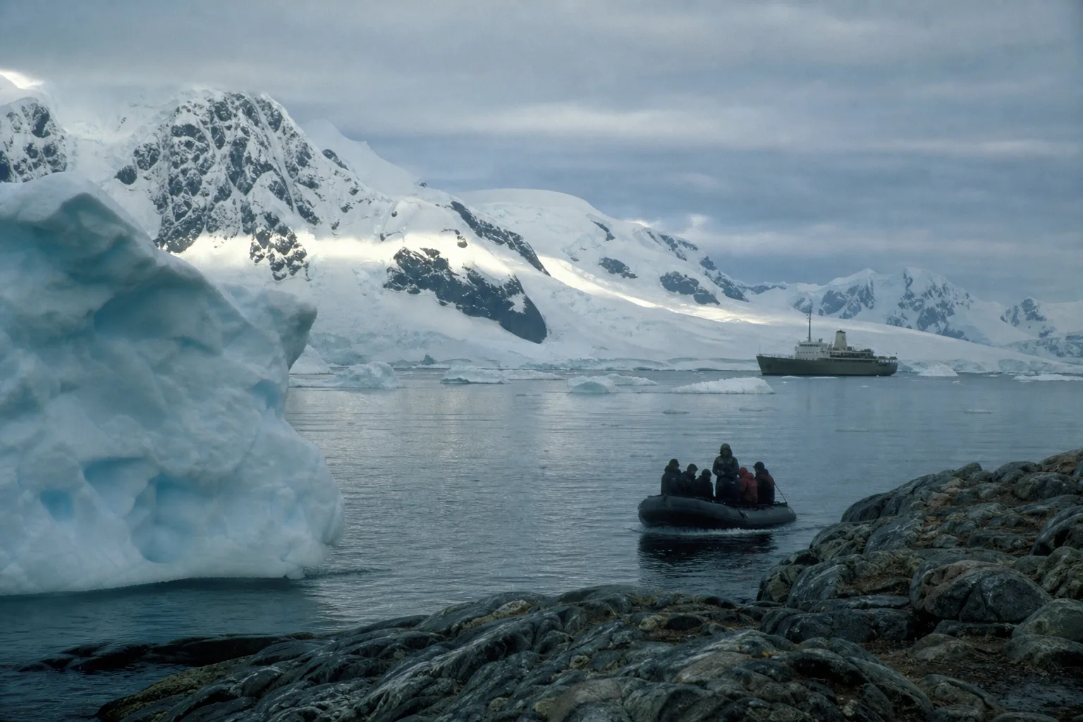 A small boat of travellers heading ashore in an Antarctic bay, a plain ship at anchor among icebergs