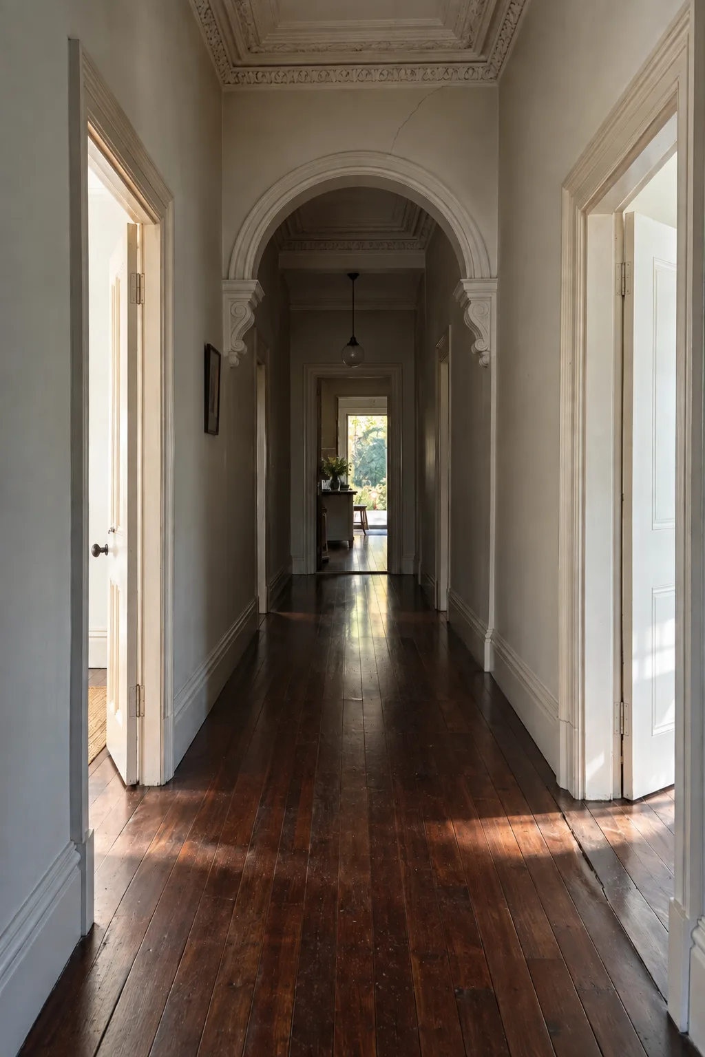 A long villa hallway with a timber arch, a fine crack above it, and afternoon light falling in two patches on the boards