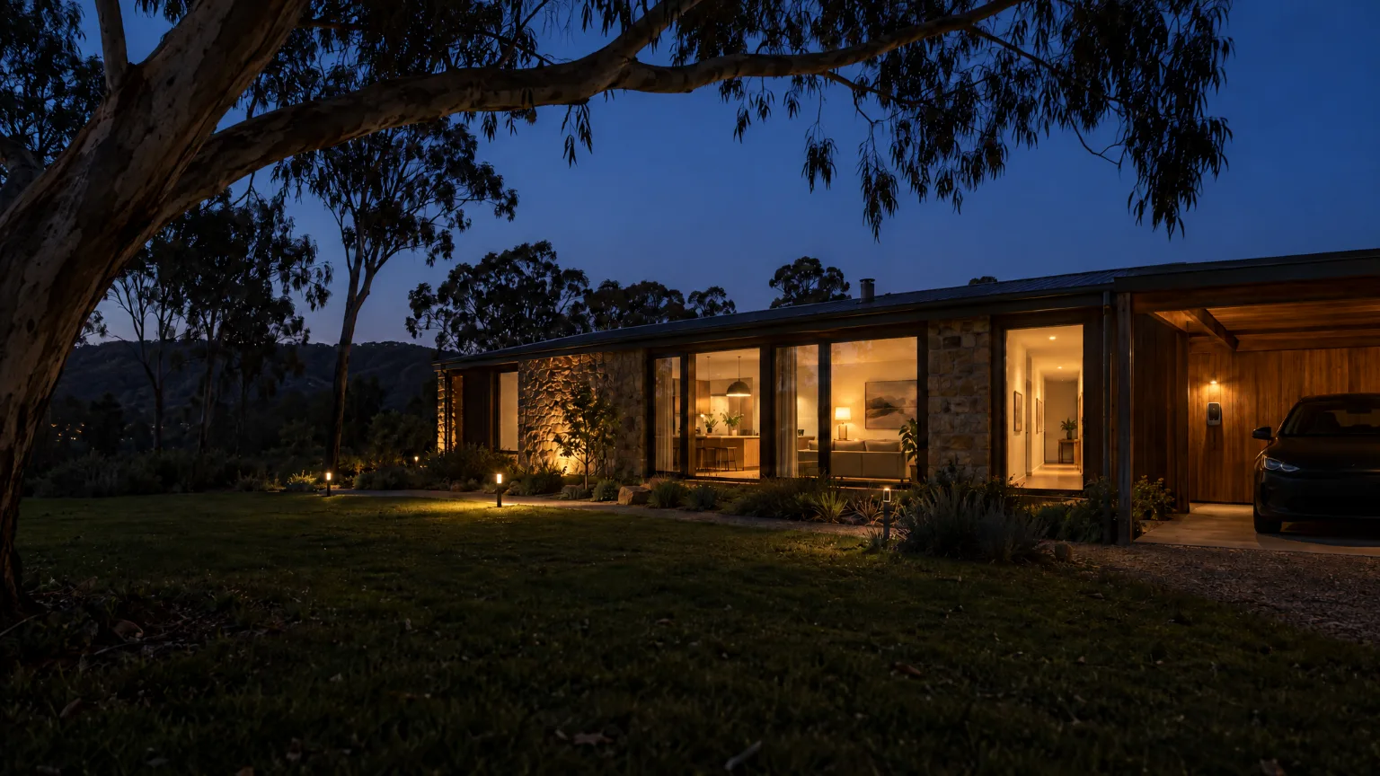 A low stone and timber house in the Hills after dark, seen across the lawn, its windows warm and a carport lit at one end