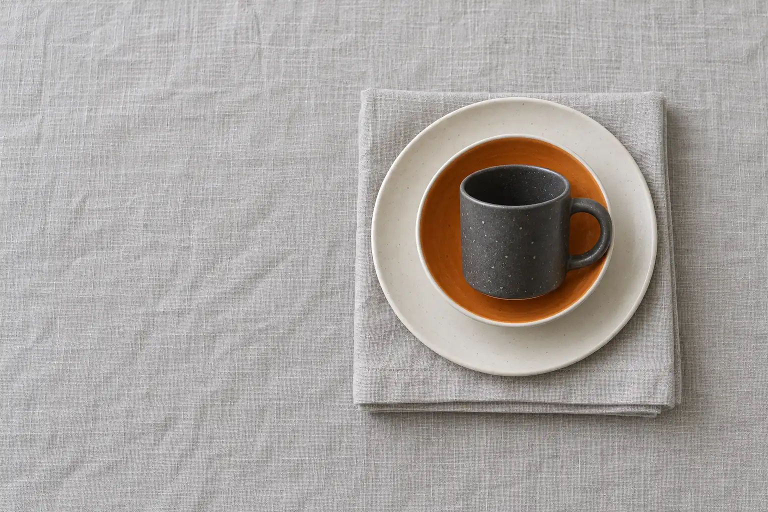 A folded linen napkin, a white side plate, a bowl glazed tangerine inside and a graphite cup, stacked one in another on a pale linen tablecloth, seen from above.