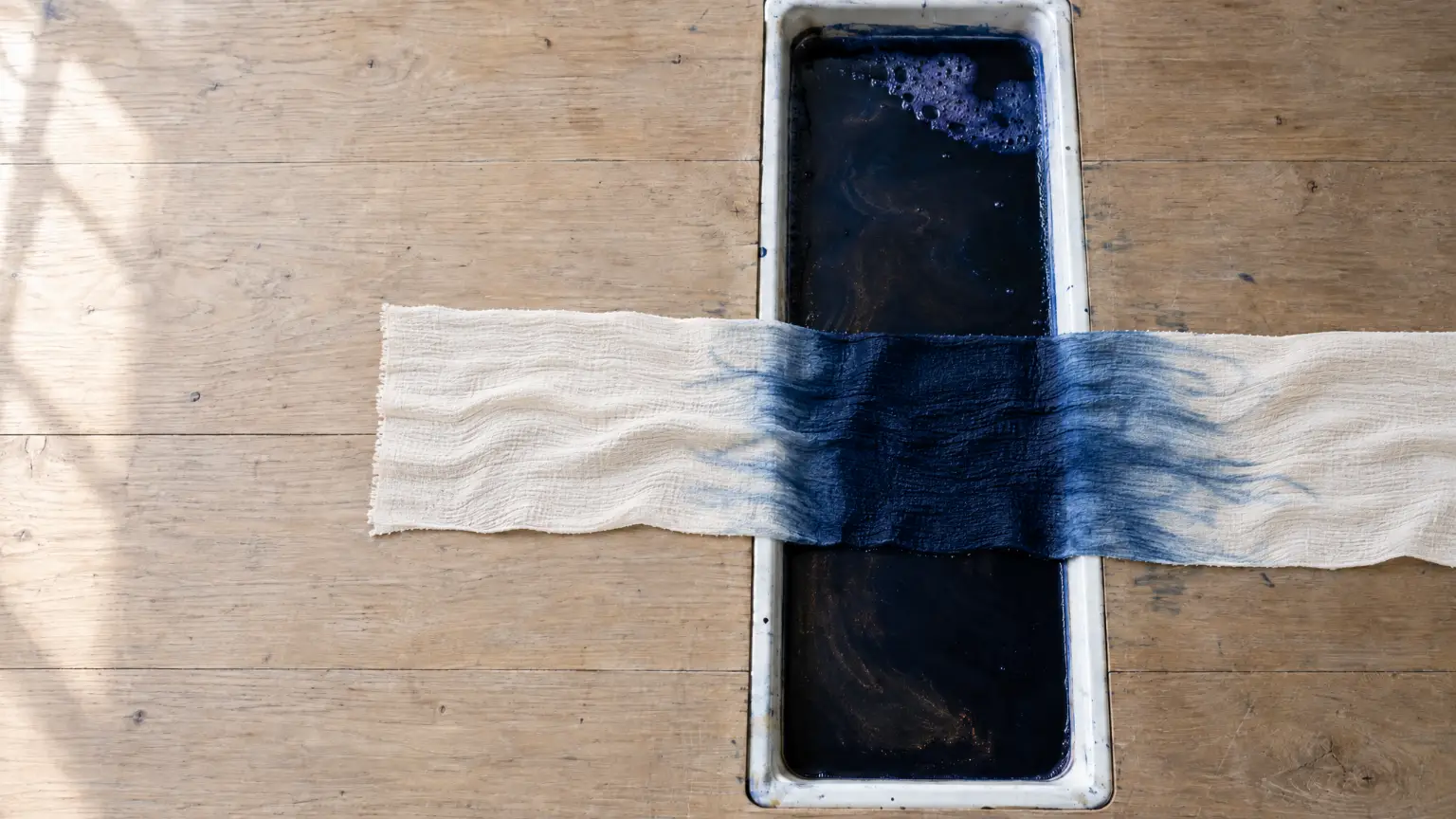 Seen from above, a length of cream crepe laid across a white enamel trough of indigo set into a pale oak table, dyed deep indigo where it crosses the dye and feathering out to cream along the cloth on either side.