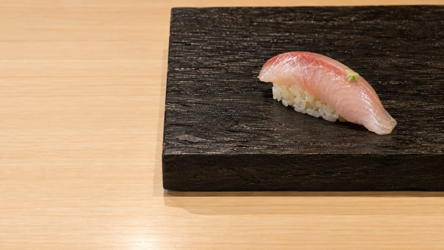 One piece of kingfish nigiri on a dark board resting on the pale hinoki counter, the empty counter to its left