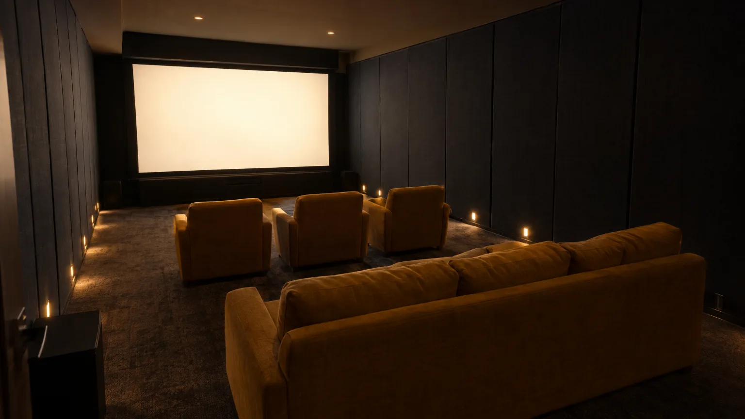 The cinema room as the lights go down: a blank screen glowing at the far end, dark fabric walls, three mustard armchairs and a mustard sofa pulled off the back wall, step lights along the skirting