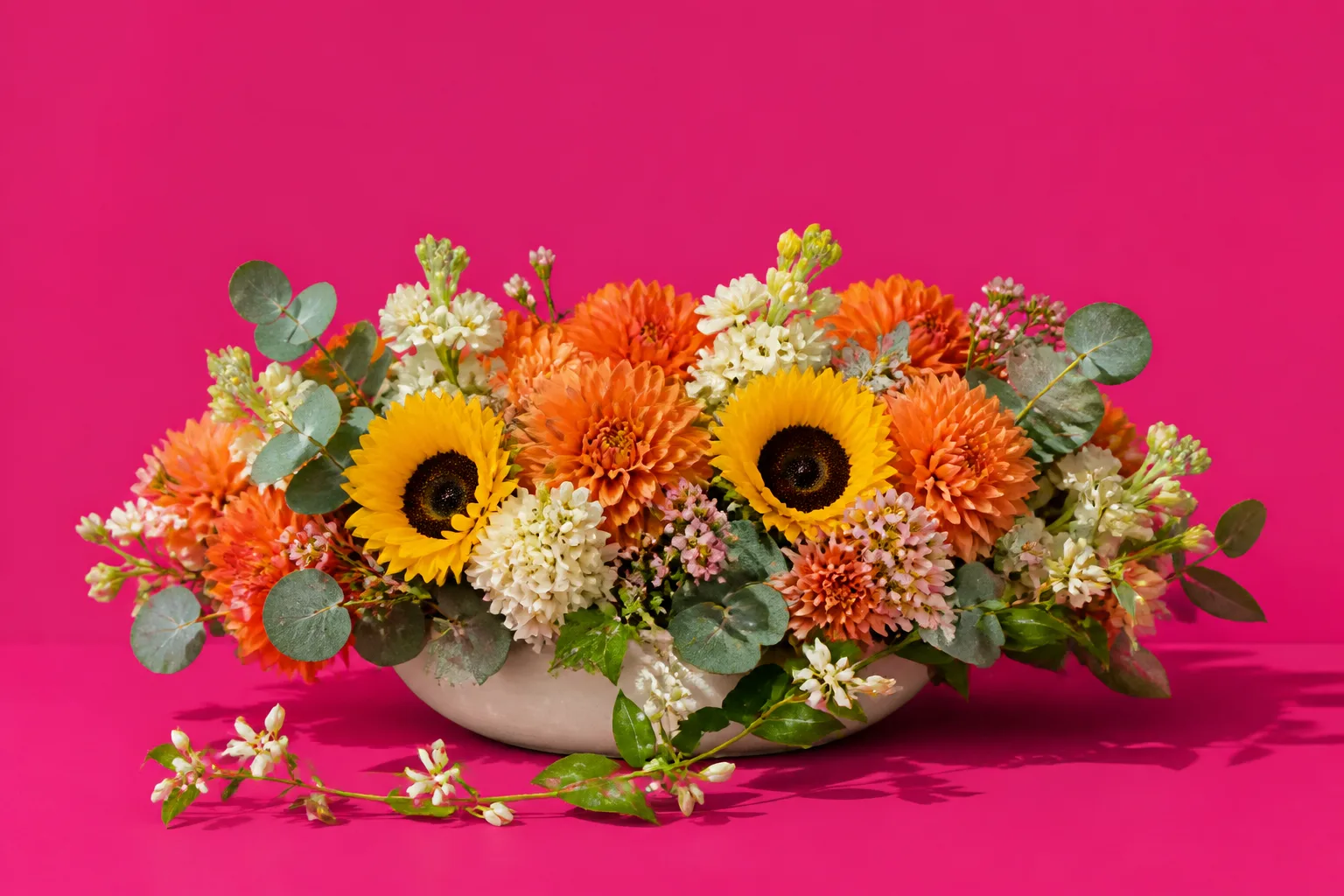 A low heaped arrangement of tangerine dahlias, sunflowers, pale pink waxflower and eucalyptus in a cream bowl on a hot magenta table, jasmine trailing over the edge