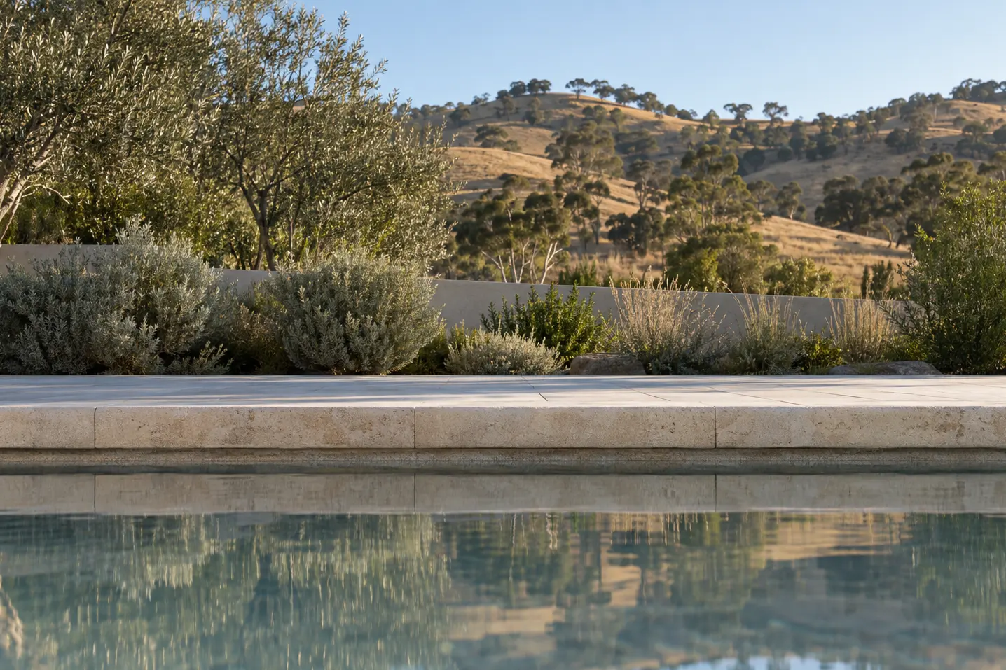 At water height across a still pool to its limestone coping, with olive trees and the dry foothills beyond.
