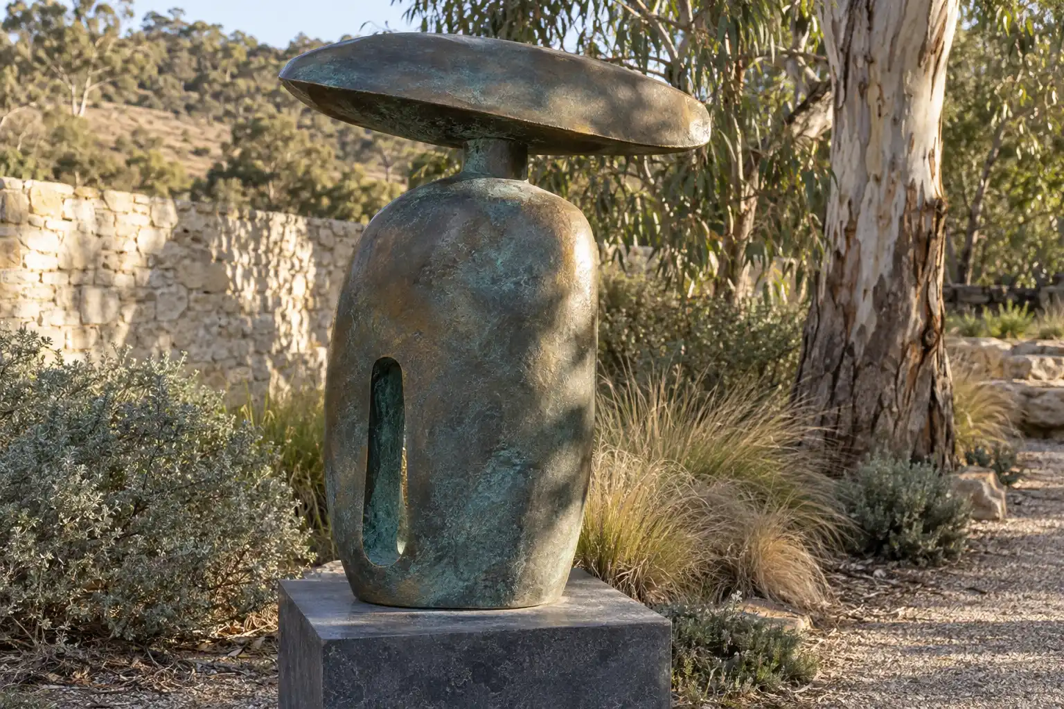 A bronze standing stone with a narrow slot and a flat capstone, blue-green in its hollows, on a bluestone plinth in a dry garden in late sun