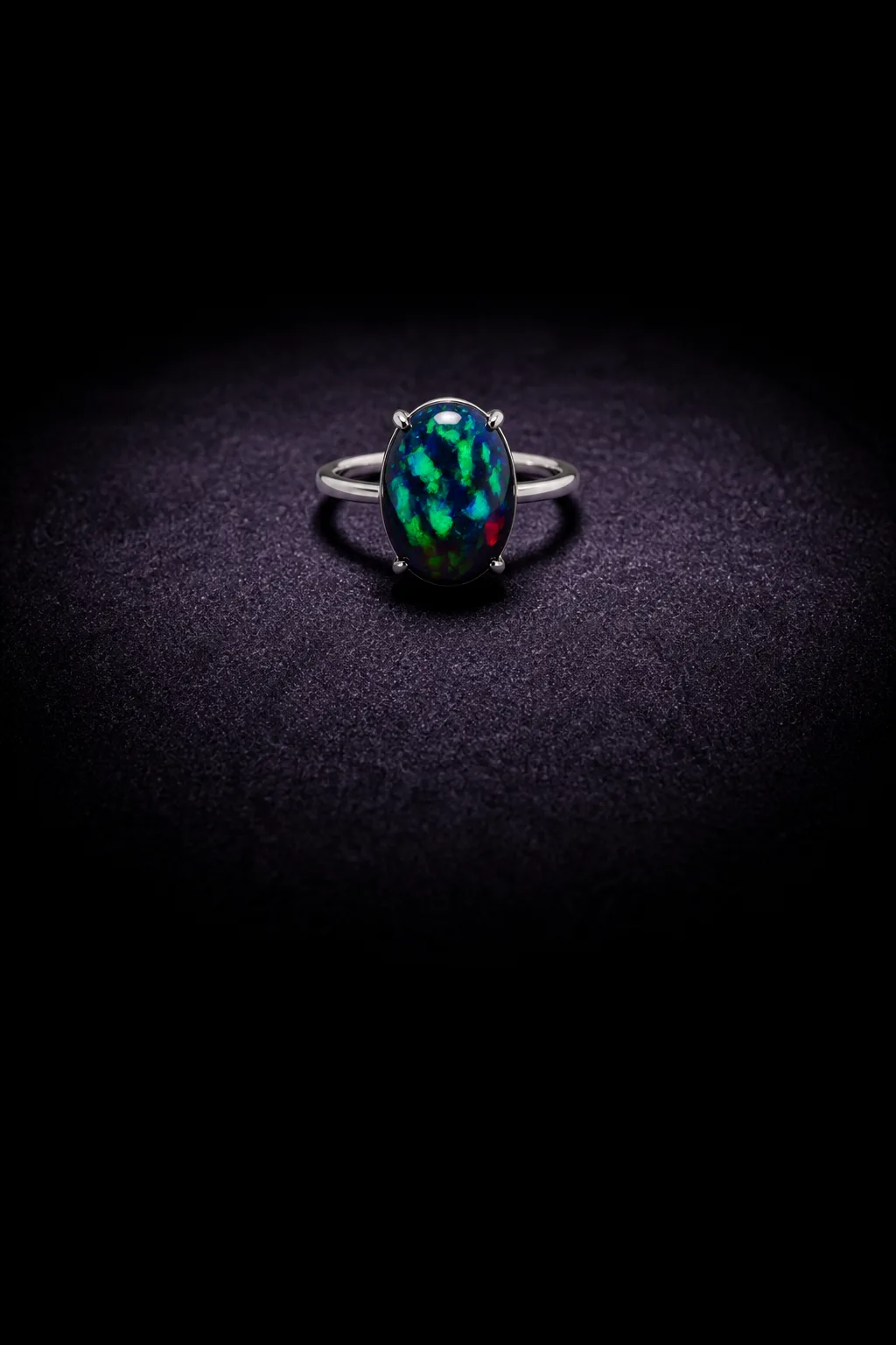 A platinum ring with an oval black opal held in four claws, lying on violet-black velvet under one overhead lamp; green and blue fire across the stone.
