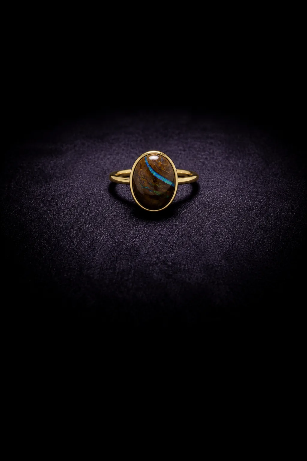 A yellow-gold ring with an oval boulder opal in a bezel on violet-black velvet; a blue vein runs through brown ironstone.