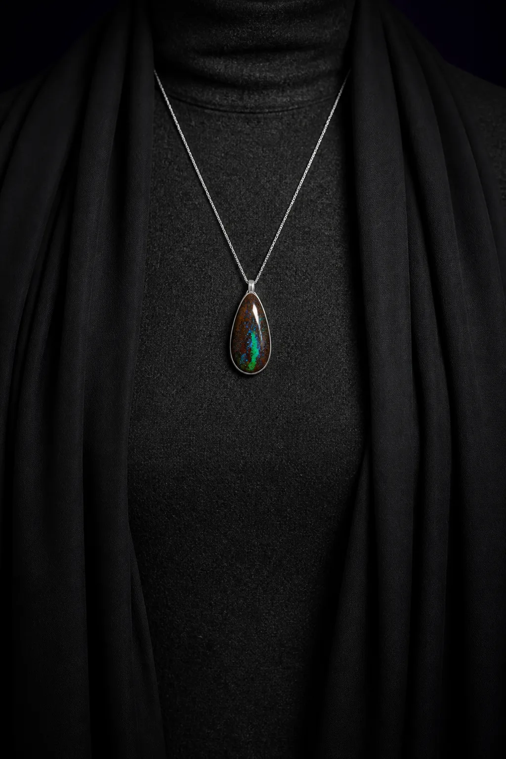 A pear of boulder opal in a platinum bezel on a fine chain, worn over a high-necked charcoal wool garment beneath a dark draped scarf.