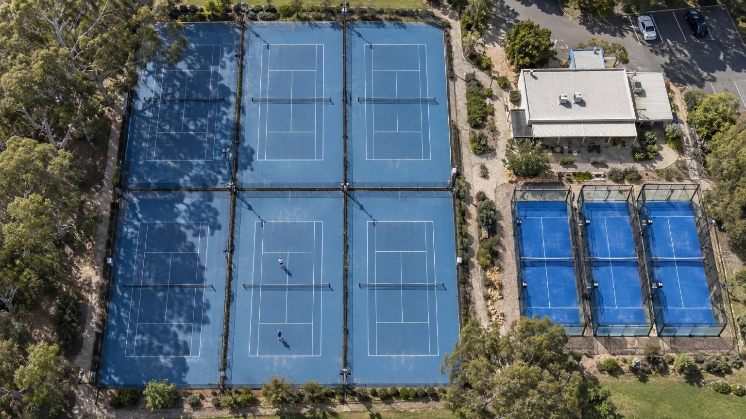 The club from above: six blue hard courts in two rows, three glass-walled padel courts beside them, a low clubhouse and gum trees shading the courts.
