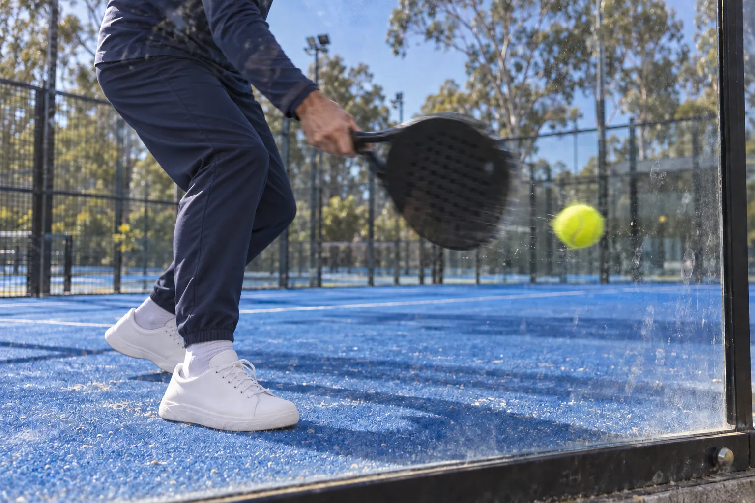Through the back glass of a padel court: a player's legs and bat mid-swing, the yellow ball just coming off the pane.