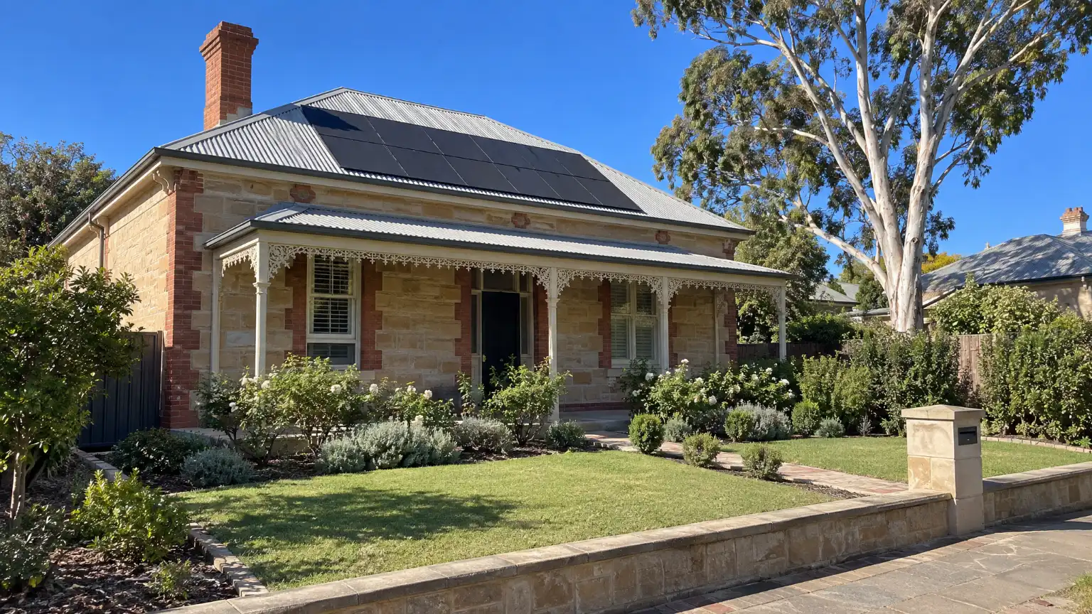 A honey-stone villa in mid-morning sun, dark panels on its hipped iron roof, the brick chimney beside them and a tall gum in the next garden