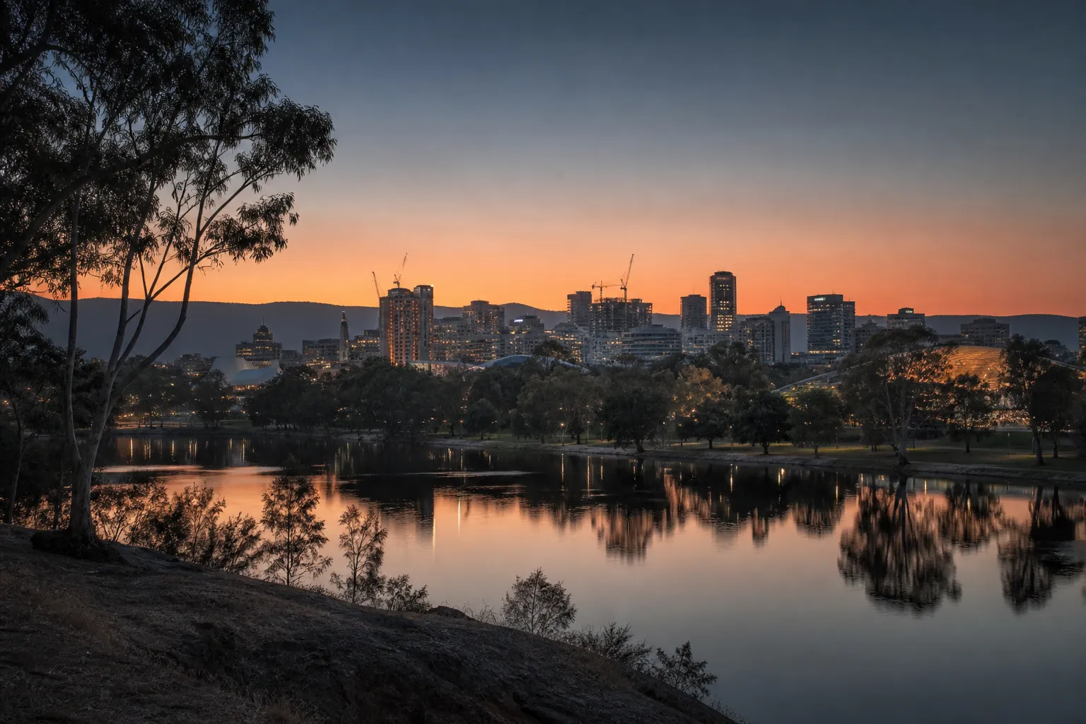 The Adelaide city skyline at dusk seen across the River Torrens, with the hills faint behind