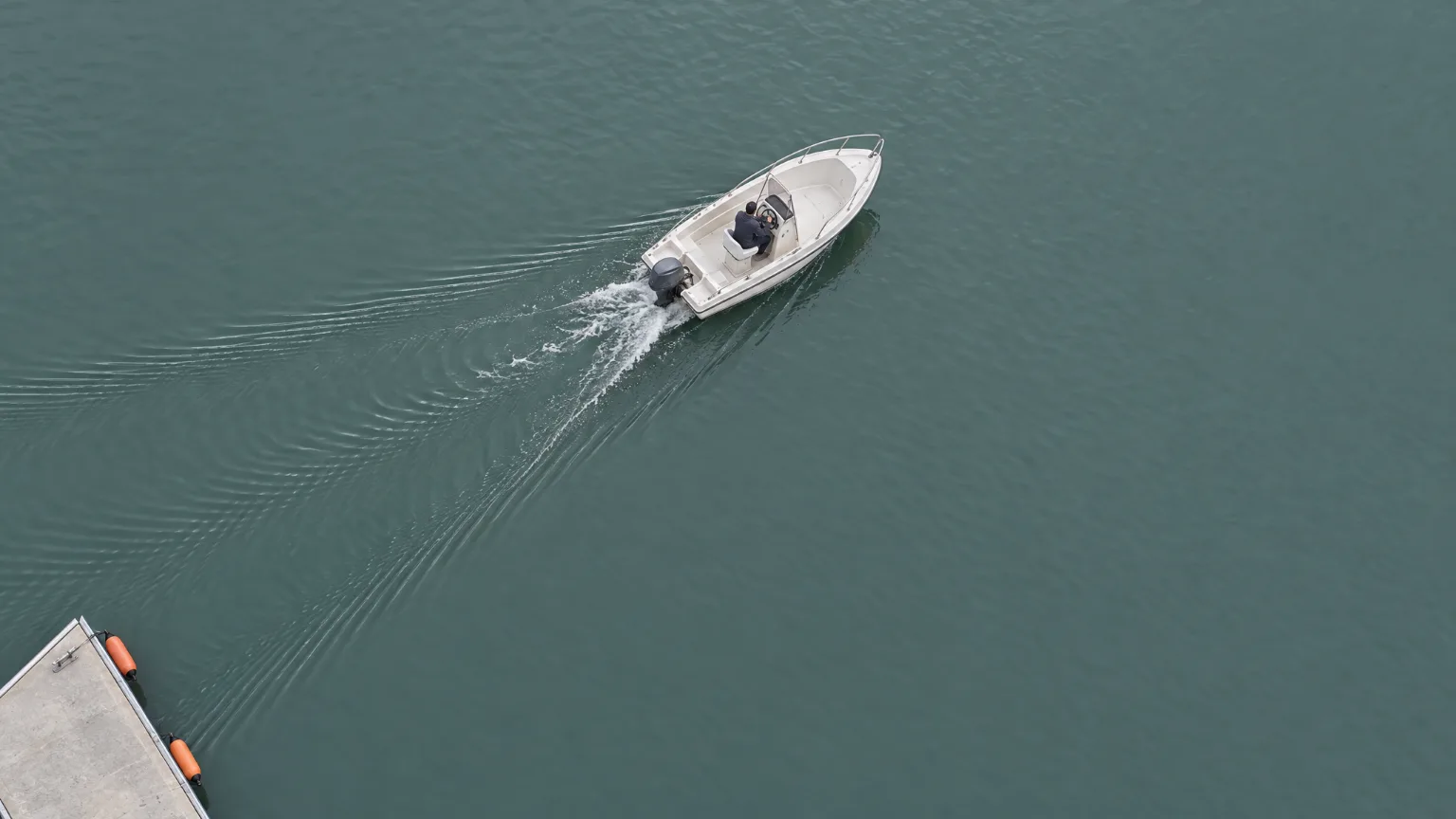 From straight above, a white runabout under way on grey-green water, its wake opening in a narrow V behind it, a strip of pontoon in the corner.