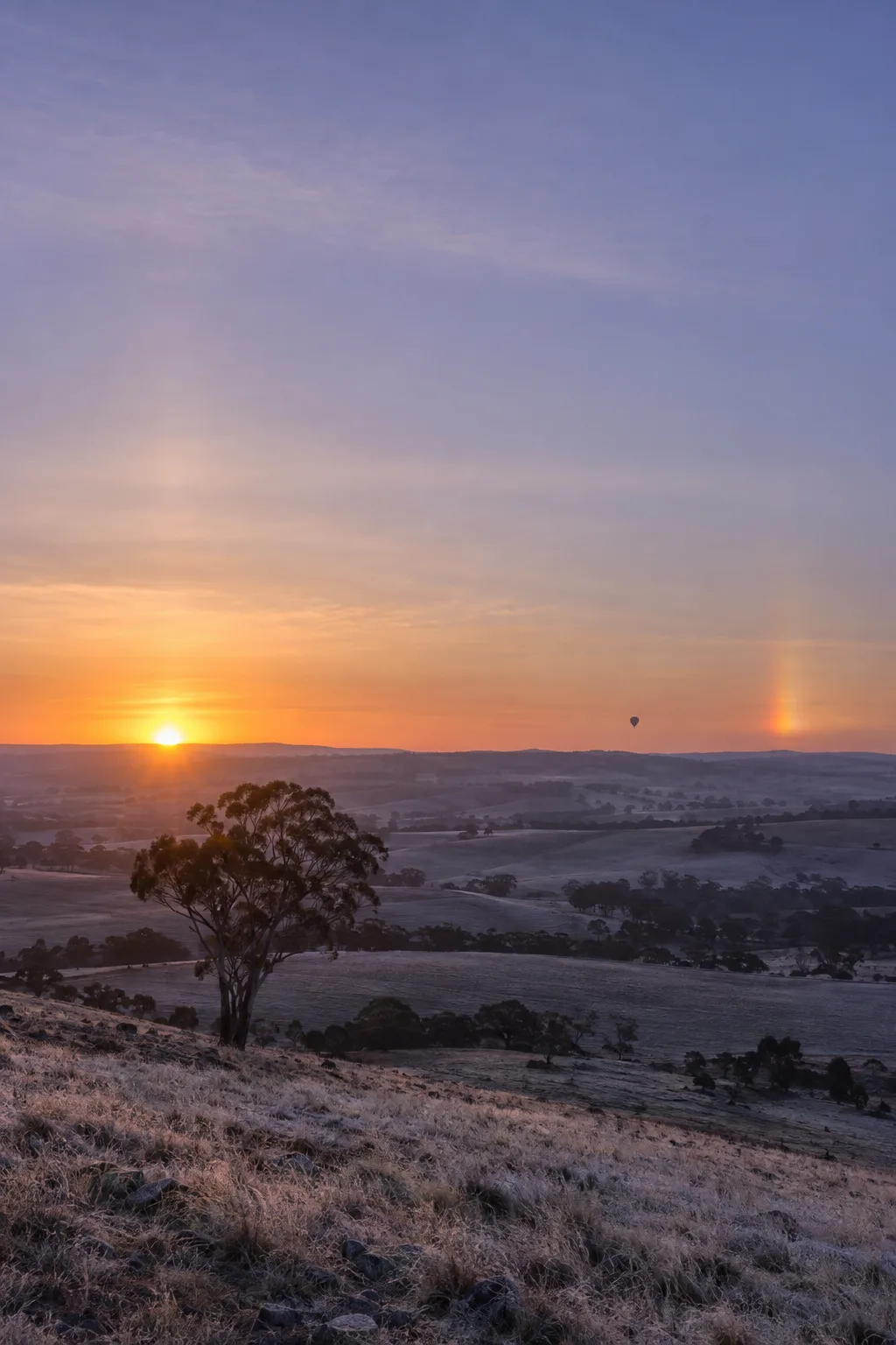 A cold dawn over the hills, a bright sun dog beside the low sun on a faint halo, a small balloon far off