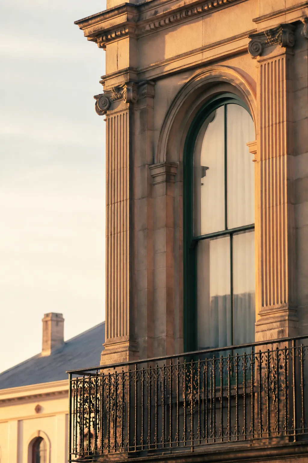 A heritage sandstone facade with an arched window, fluted pilasters and an iron balcony in low morning light