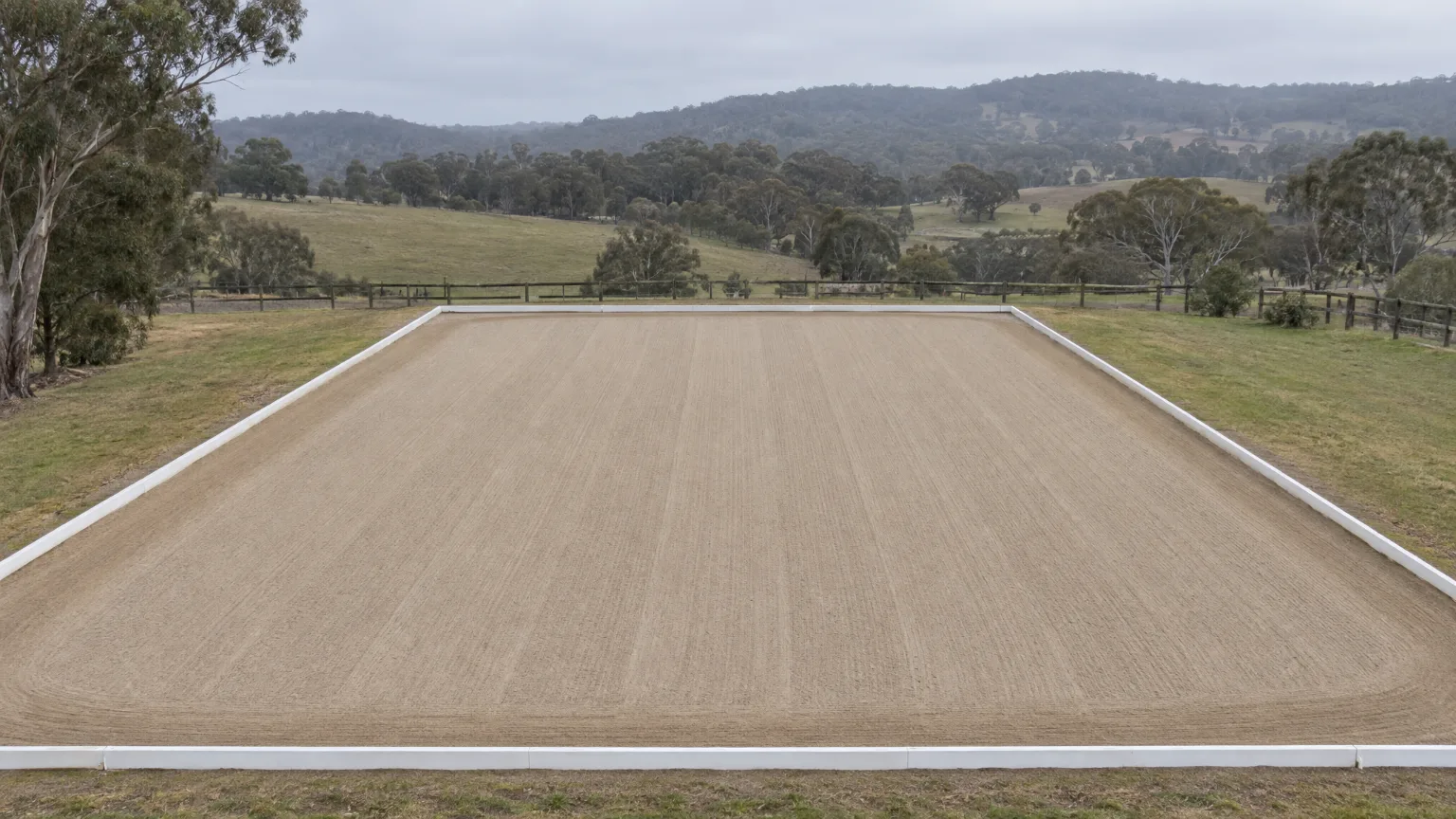 A freshly harrowed sand arena seen from above its near end, white boards running away under a grey sky towards wooded hills, a circle of hoofprints pressed in at C and a horse and rider halted beside it.