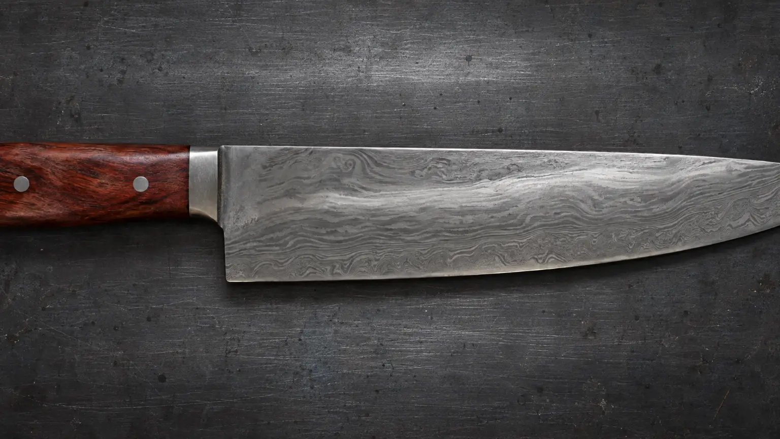 A pattern-welded chef's knife with a red gum handle, lying across a dark grinder-scratched steel bench, seen from directly above.