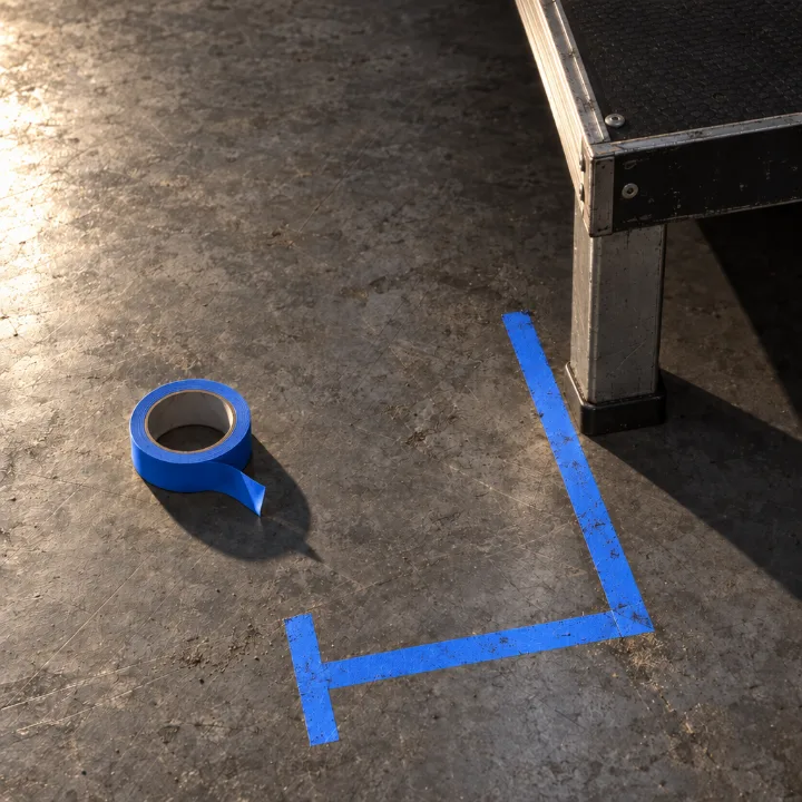 A corner mark in blue tape on the grey floor, the roll beside it and the edge of a stage deck.