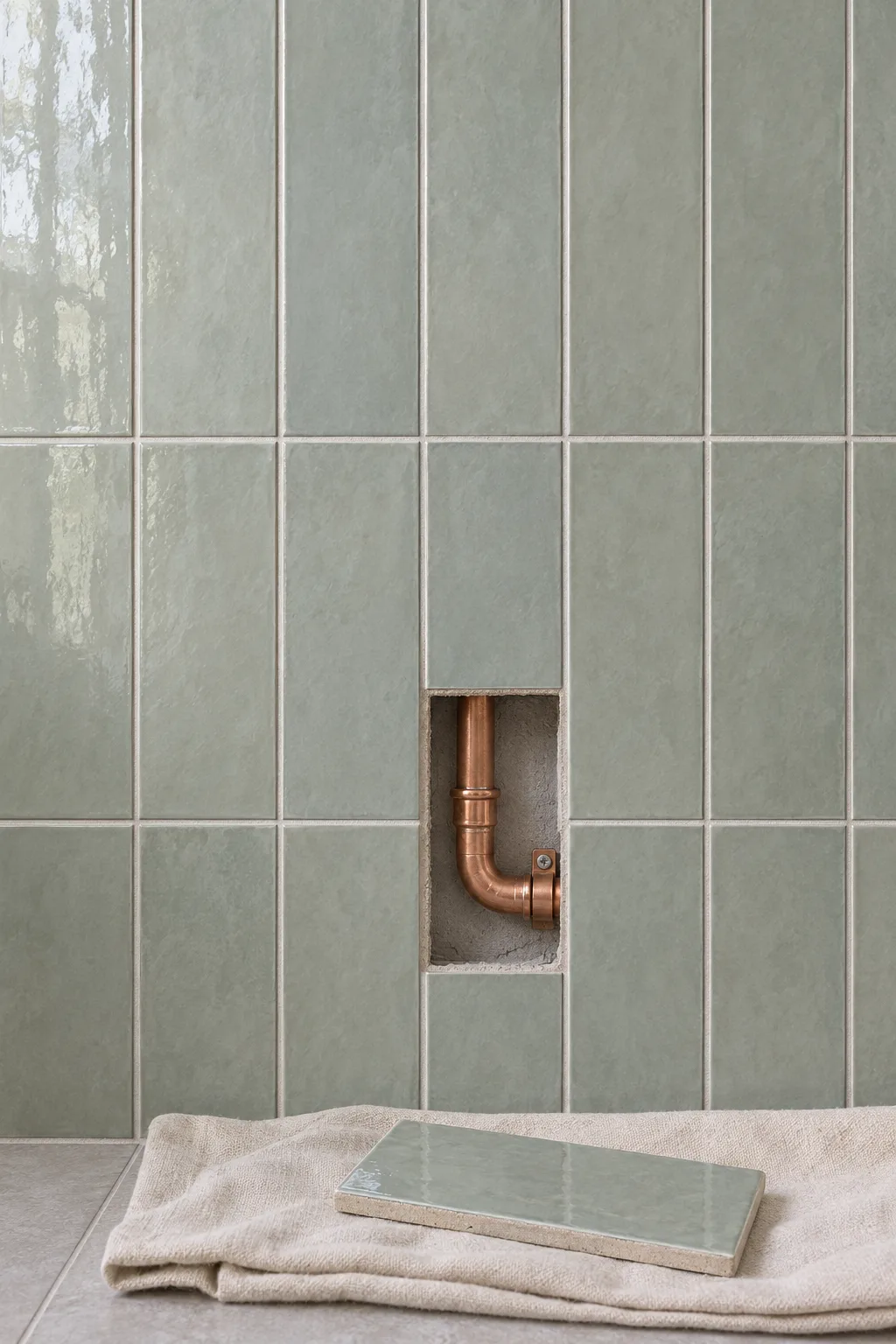 One tile lifted out of a tiled wall, showing a short run of copper pipe with a new joint behind it; the tile rests on a drop cloth below.