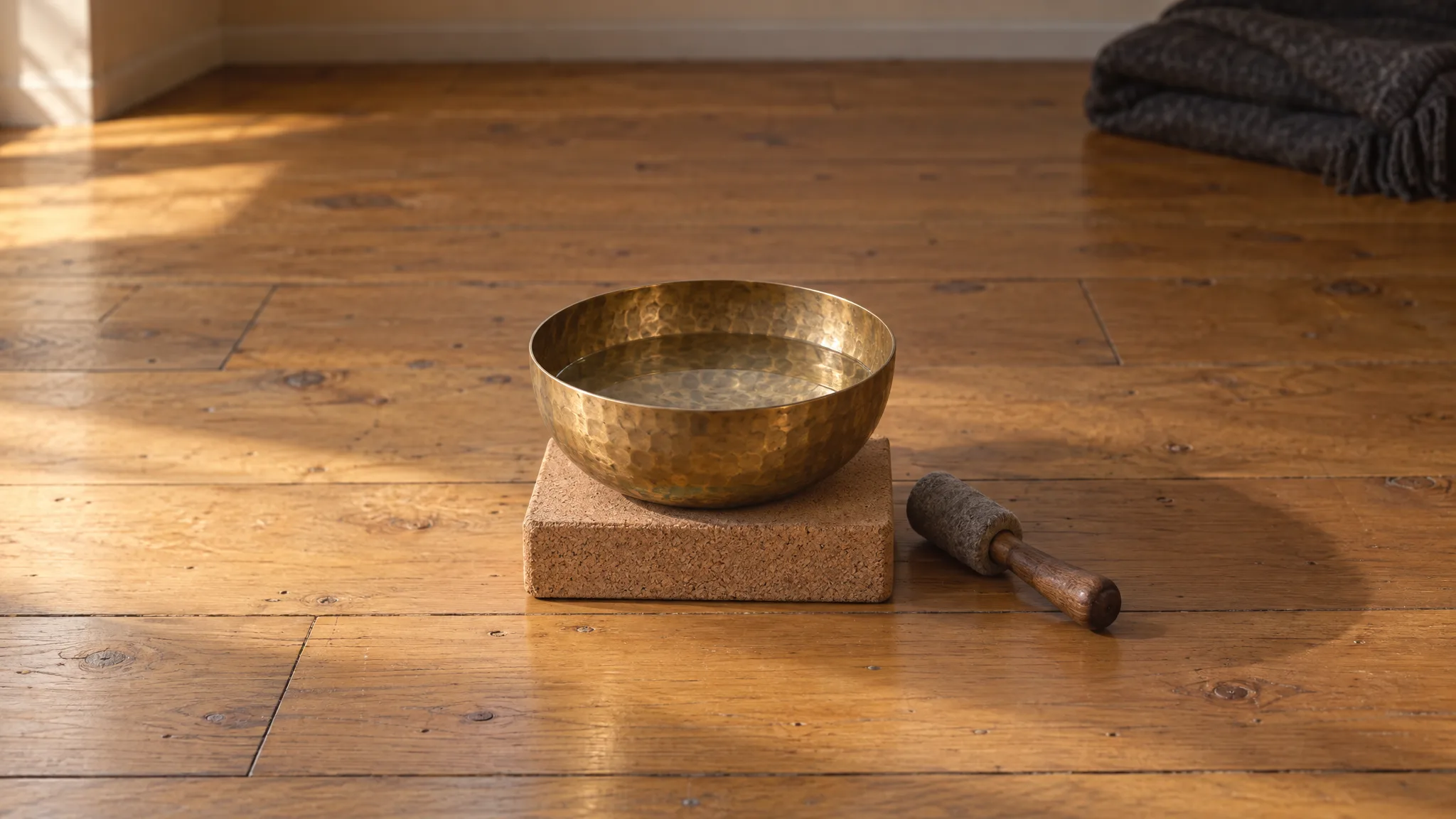 A hammered brass bowl holding a little still water, on a cork block on a timber floor, a felt-headed striker beside it.