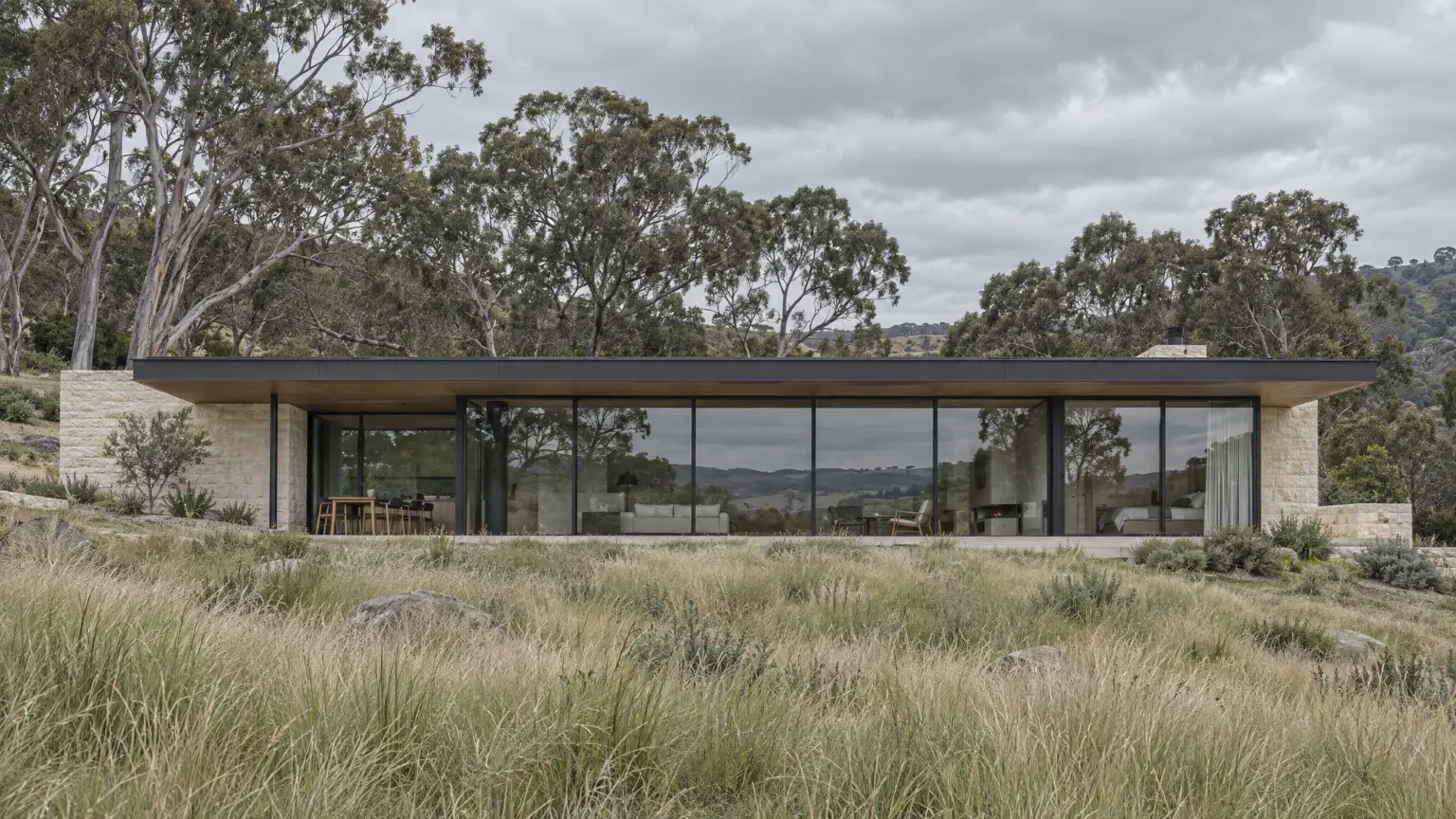 A long limestone house set into a grassy slope in the hills, its glass front reflecting a grey sky.
