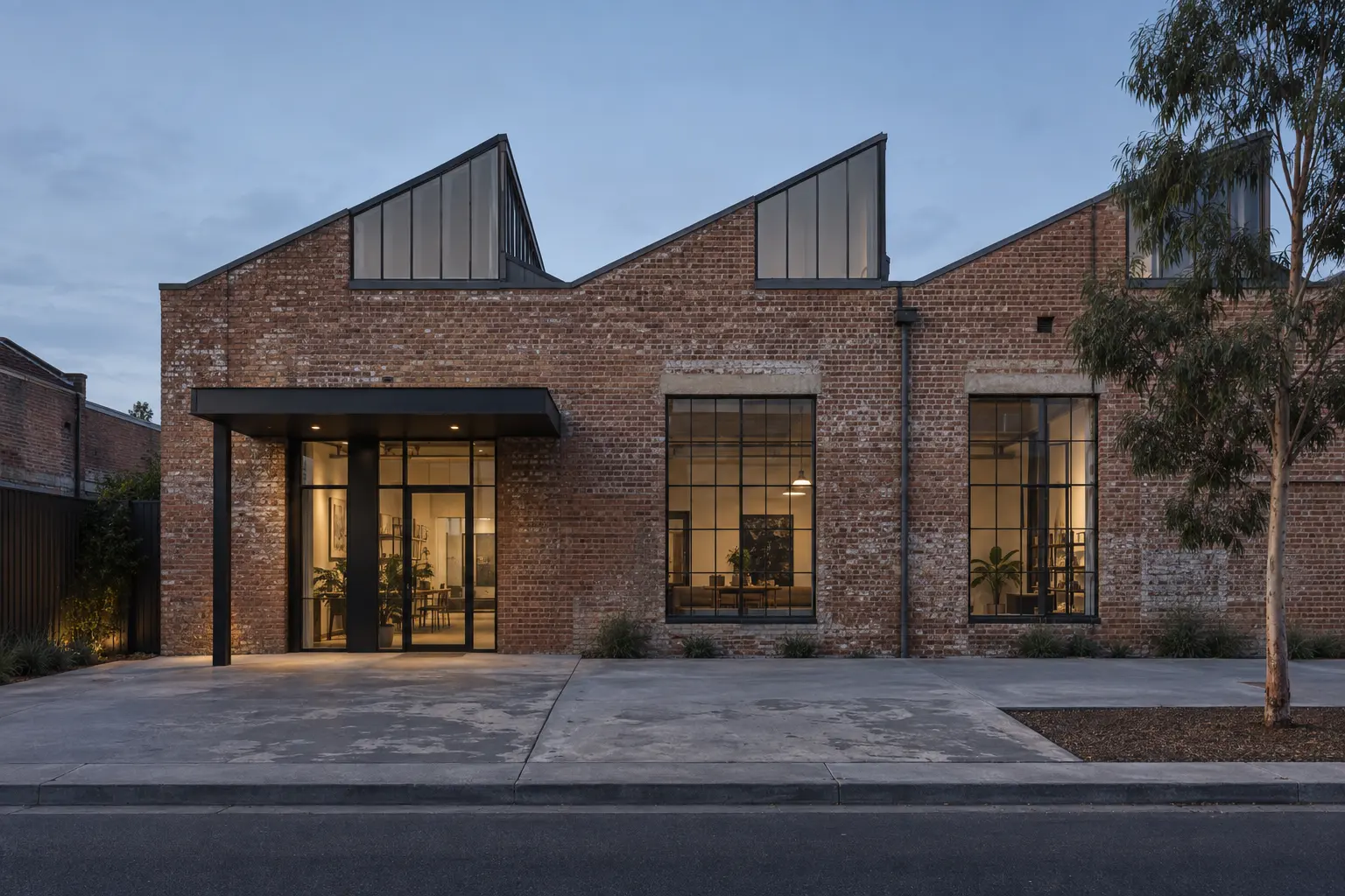 A brick warehouse with a sawtooth roof at dusk, warm light in its tall steel windows.