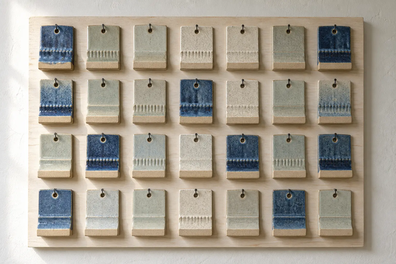 Rows of small glaze test tiles hanging on a timber board: deep blue breaking to tan, a pale grey-green wash and a speckled white
