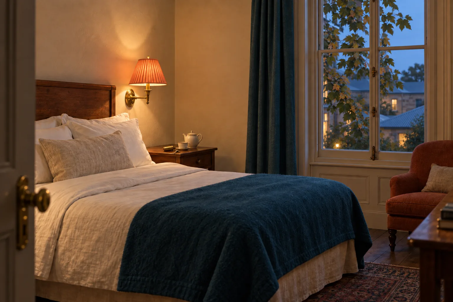 A Courtyard room with the lamps on: a bed made in heavy linen, a coral-shaded sconce, and plane-tree leaves close to the window.