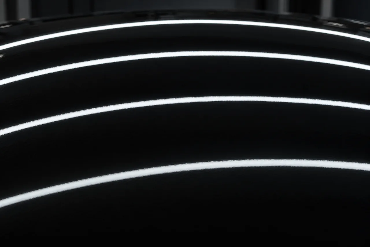 The crown of a gloss black bonnet after polishing, overhead strip lights reflected across it as long, sharp curving bands.