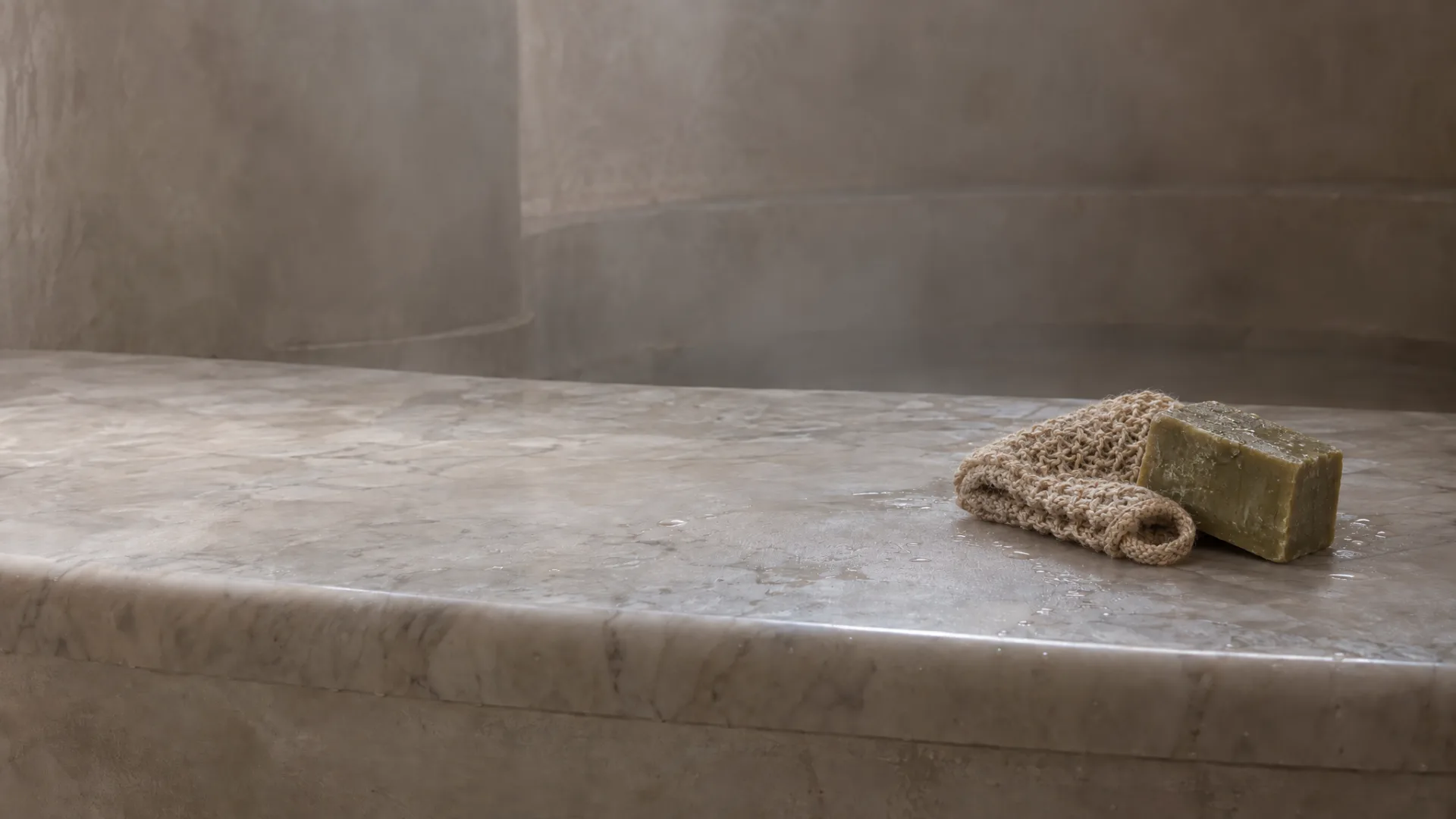 A heated white-grey marble slab in the hot room, a woven mitt and a block of olive-oil soap near its far end