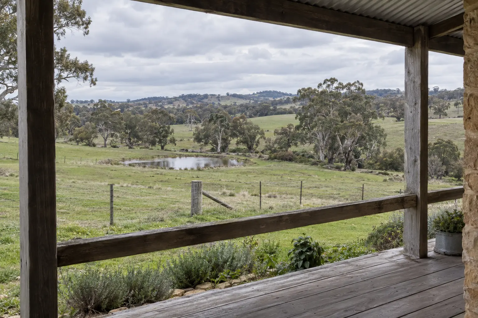 From a stone cottage's verandah, looking between two timber posts over green paddocks to a dam and a line of red gums.