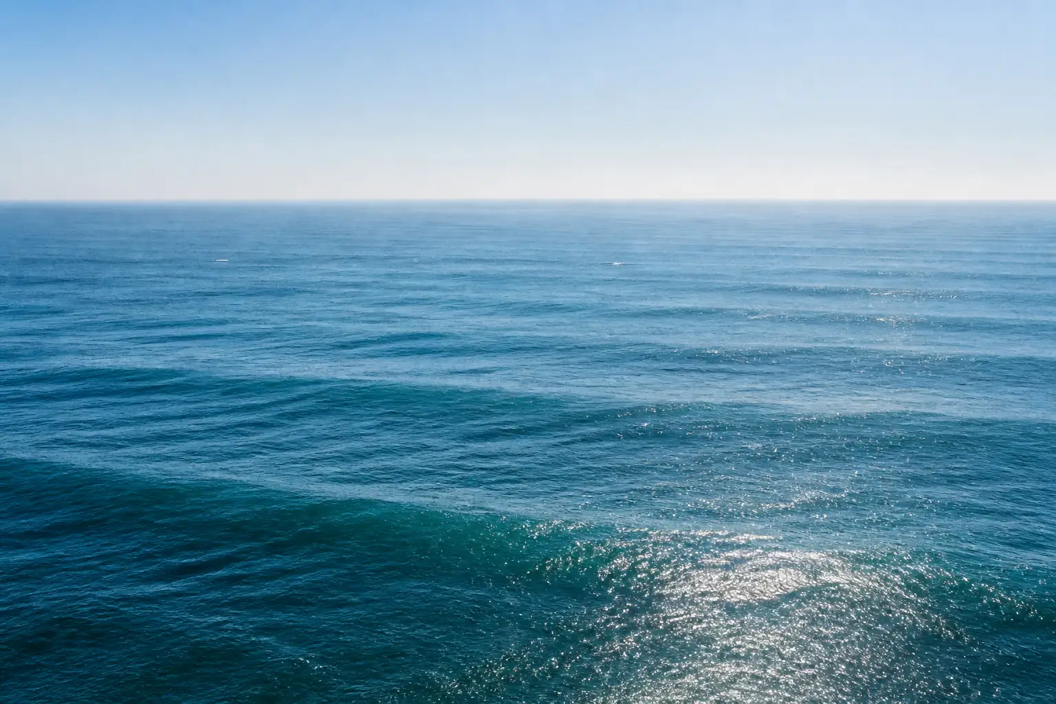 Open sea by day: a long swell running towards you under a pale sky, a breeze feathering the water and sunlight scattered across it.