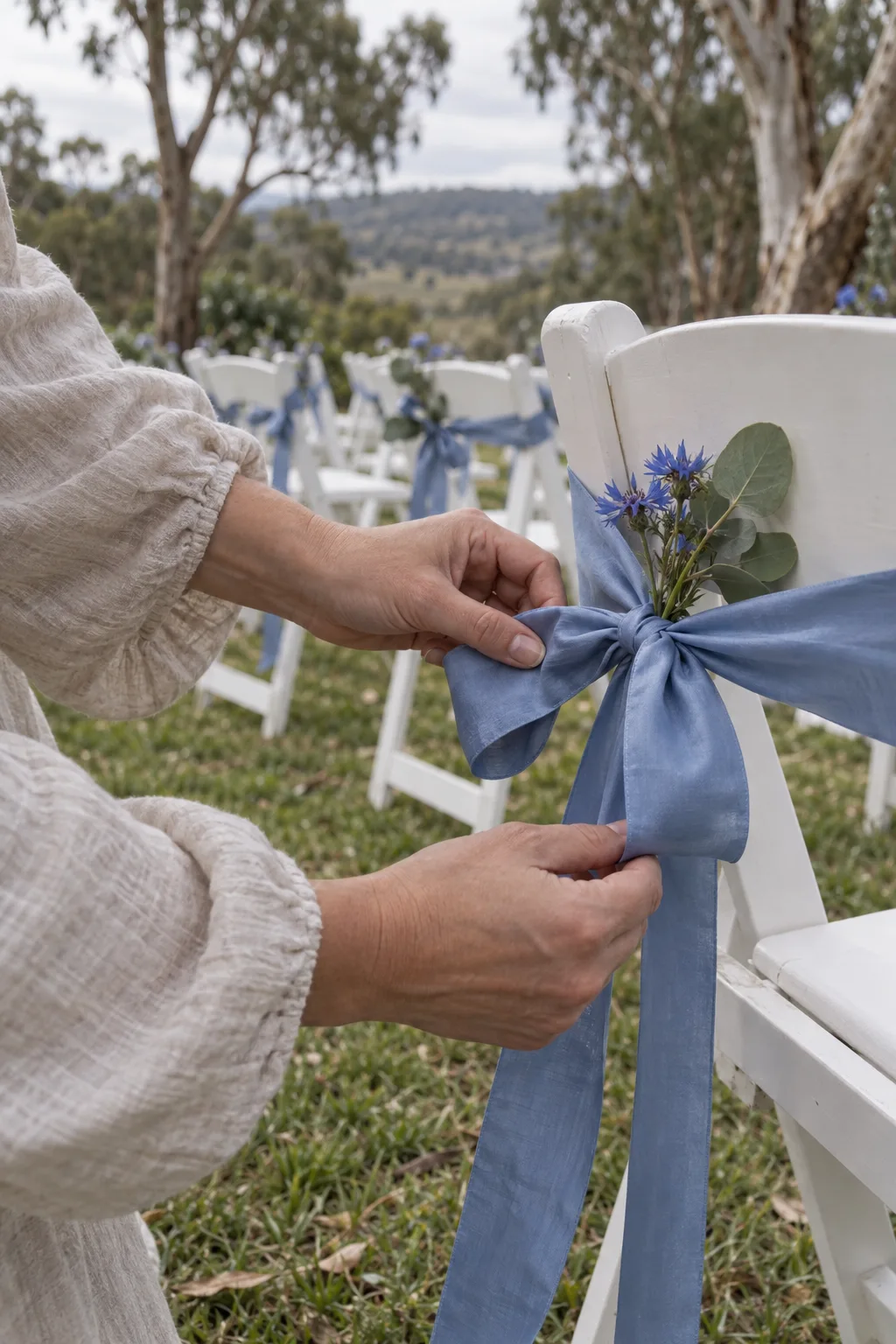 Hands tying a cornflower-blue ribbon onto the back of a white garden chair.