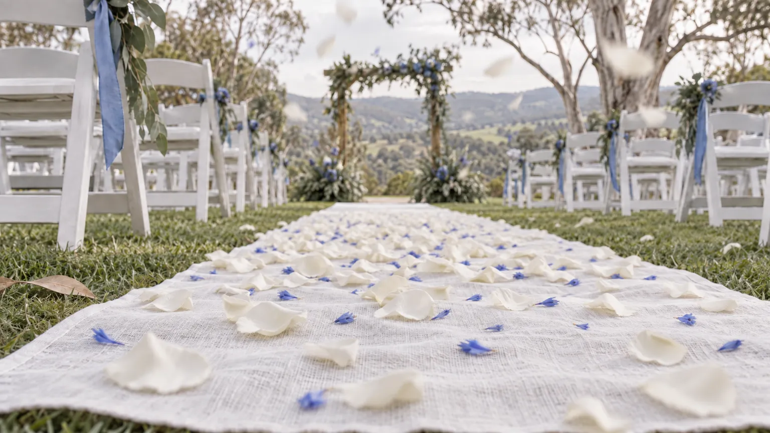 Down at the white linen runner of a garden ceremony aisle in the Adelaide Hills, white and cornflower petals strewn across it and a few still falling, the chairs soft on either side and an arbour of gum leaves and cornflowers far off.