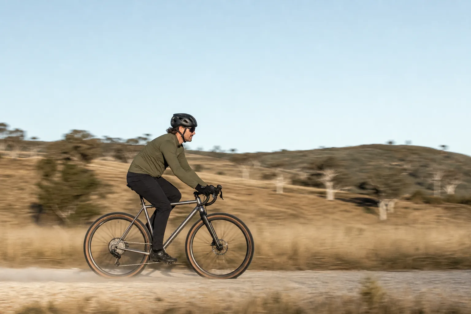 A rider in a long-sleeved olive jersey and helmet on a bare titanium gravel bike, side-on on a pale gravel road in the Adelaide Hills, the grass behind streaked by the pan.