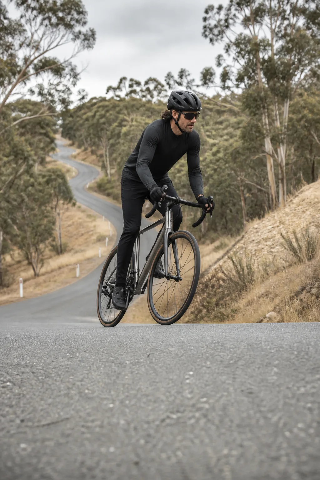A rider in long sleeves and a helmet climbs a winding sealed road between gum trees on a bare titanium road bike.