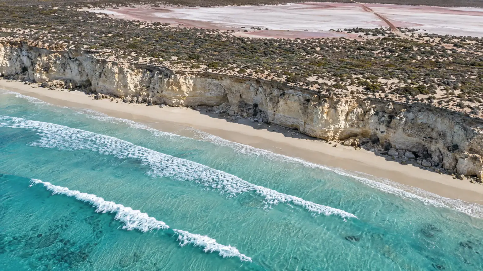 Looking steeply down at the gulf coast: three lines of surf, a narrow beach under a pale limestone cliff, and a salt pan beyond the dunes