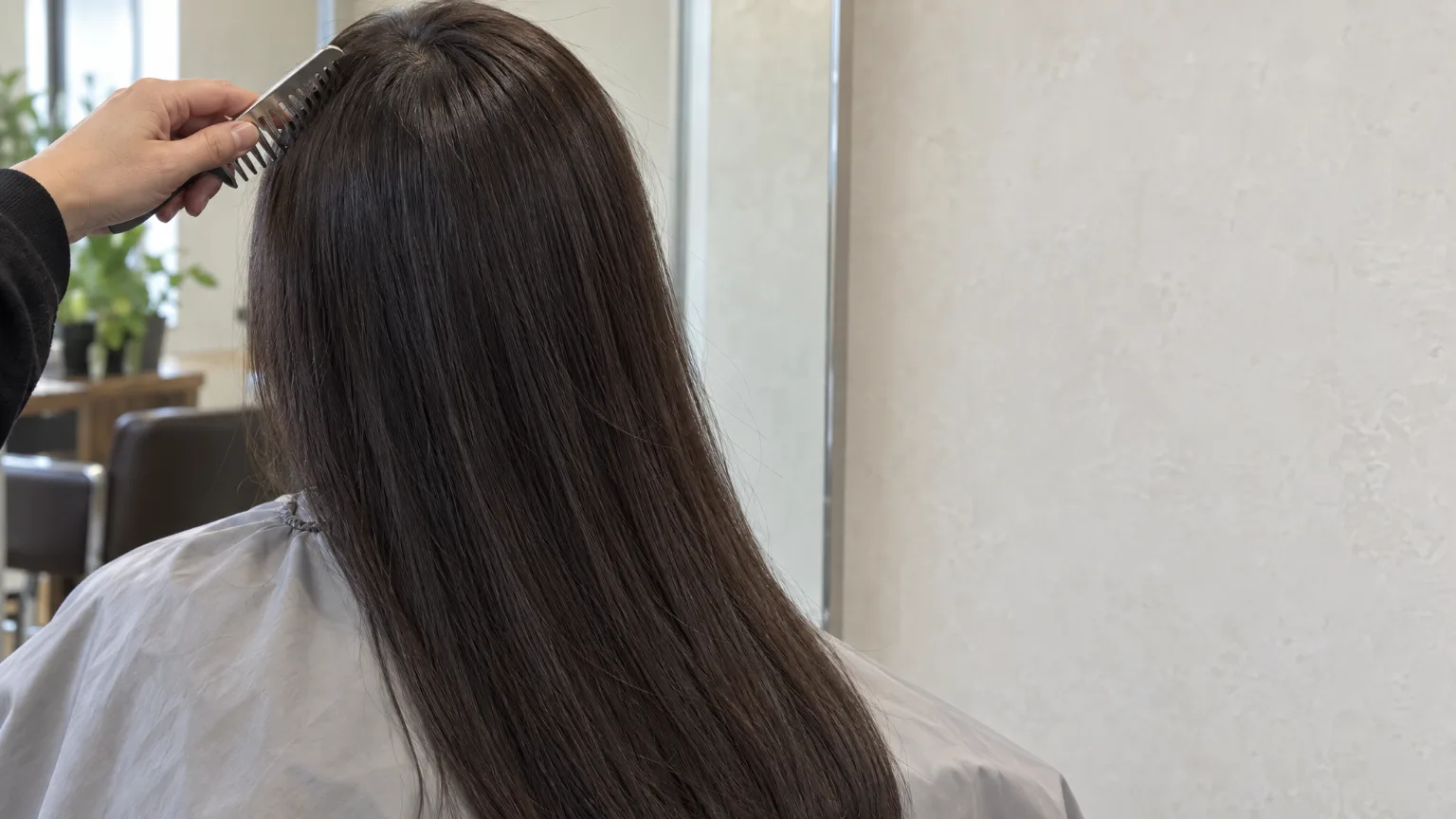 Long dark hair seen from behind over a pale salon cape, a stylist's steel comb at the crown, the plaster wall and a mirror's edge beside it